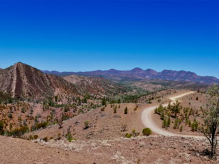 A winding dirt road through a dry, rocky landscape with sparse vegetation and distant mountains under a clear blue sky.